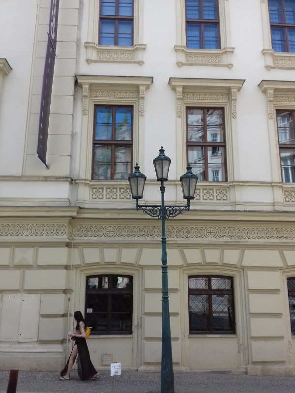 Beautiful woman with long black hair wearing long black leg-revealing dress, walking past old and grand cream-colour hotel. Old-fashioned ornate cast-iron street light, which forks into three lamps, is also on the pavement in front of the building.